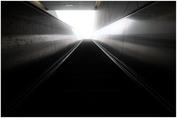Blick nach oben auf eine Rolltreppe, die in helles Licht führt, mit reflektierenden Metallwänden an den Seiten.