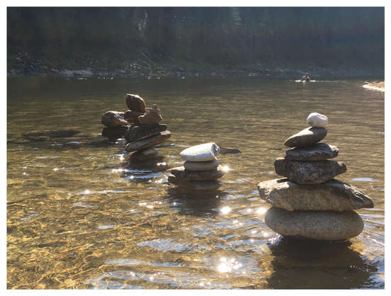 Photo of several stacks of balancing stones in a river: many of them smooth, wide and flat closer to the base, tapering to smaller rounded pieces at the top. Sunlight glitters on the golden waves. In the distance are two swimmers, and beyond, a far bank lined with conifers.