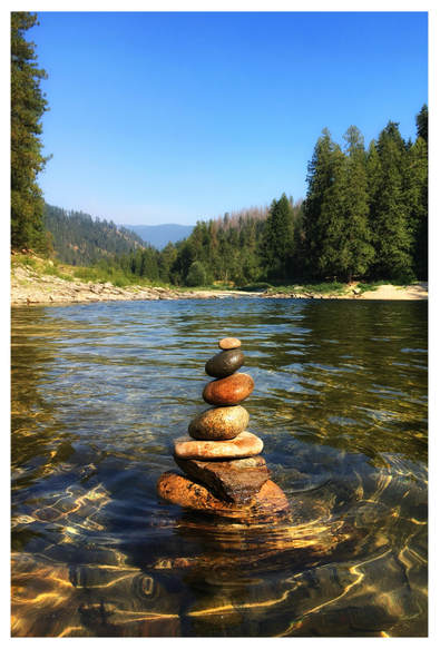 Photo of a pile of carefully balanced stones in the middle of a river. The stones are wider and flatter at the base, smaller and rounder at the top. Ripples radiate out from the pile and cast sunlit patterns on the rocky riverbed, which can be seen through the clear water. A beach lies in the distance, with rocks and sand eventually giving way to a dense conifer forest. Further off are more forested hills, fading into blue under a clear blue sky.