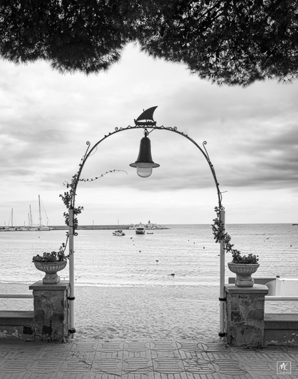 Black and white photo looking through a thin metal decorative archway leading from a paved seaside walkway to a beach and the Mediterranean Sea beyond. 