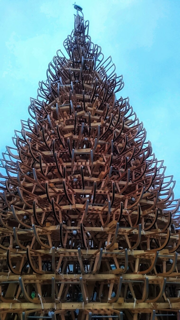 A striking, large-scale outdoor sculpture resembling a towering tree, meticulously constructed from stacked wooden sledges. The sledges are arranged in concentric layers, forming a conical shape that ascends high into the sky. Each sledge is carefully positioned with the curved fronts facing outward, mimicking the branches of a tree and creating a sense of intricate texture and depth. The warm, natural tones of the wooden sledges contrast beautifully with the pale blue sky in the background, enhancing the visual impact of the installation. The sculpture is both whimsical and impressive, transforming everyday winter objects into a captivating work of art.