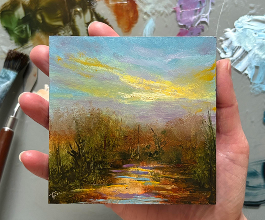 Photo of hand holding a small 4"x4" original autumn landscape oil painting by Tisha Mark. The painting shows a body of water reflecting colors from a sunset sky surrounded by trees that are turning red, brown, and gold. A paint palette with puddles of oil paint and a paintbrush and palette knife are visible in the background.
