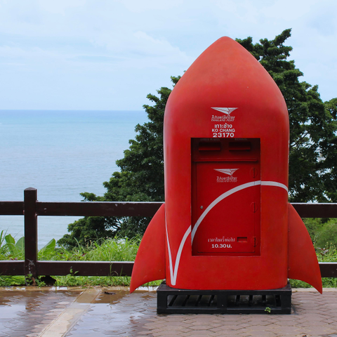 das qudratischebild zeigt einen roten briefkasten in raketenform mit seitlichen flügeln der thailändischen post. er steht auf einem gepflasterten bereich, der nass aussieht. im hintergrund sieht man einen holzzaun, das meer und bäume. auf dem briefkasten steht schrift in thailändisch und englisch.