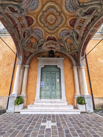 The entrance of a historic church, featuring an ornate architectural style. The doorway is framed by two classical columns supporting an intricately decorated archway. The arch is adorned with elaborate frescoes in warm tones of gold, red, and green, showcasing floral and geometric patterns.

The door itself is painted a muted green and has a traditional double-door design with panels. Above the door, a small sign is visible. Flanking the entrance are two potted plants, adding a touch of greenery to the scene. The floor is paved with rectangular stone tiles, and a small cross symbol is embedded in the pavement directly in front of the entrance. The walls of the building are painted in a soft ochre colour, enhancing the historic and artistic ambiance of the setting.