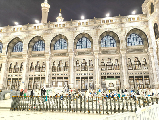 Crowds of pilgrims sit and walk outside the illuminated Masjid-Al-Haram (The Sacred Mosque in Mecca, Saudi Arabia, at night. The mosque is a large, ornate building with a minaret, arches, and many windows.