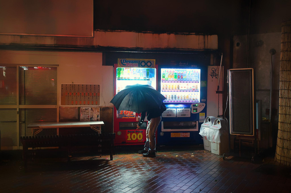 On a rainy night in Okayama, Tokyo, a man with an buys a drink from a vending machine.