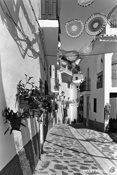 Black and white photo of a narrow, sunlit street in Guájar-Fondón, lined with white buildings. Decorative woven parasols cast intricate shadows as potted plants hang from the walls above a patterned stone walkway.