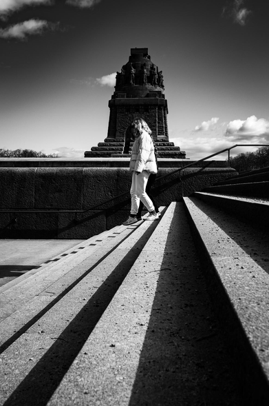A striking black and white photo shows a person walking down wide stone steps, creating bold, parallel lines that draw the eye upward. Sharp sunlight casts long shadows, adding to the graphic quality. The figure, a young woman in light clothing, is silhouetted against the light, making her a secondary element. Behind her, a massive stone monument rises into the sky. Dark and imposing, it contrasts with the bright sky, its details obscured by distance and backlighting.