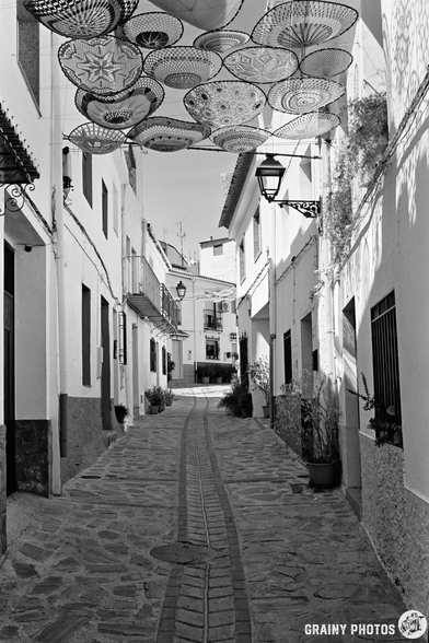 A narrow cobblestone street in Guájar-Fondón, a Mediterranean village, with white buildings on either side and decorative woven discs hanging overhead, creating shade and patterns.
