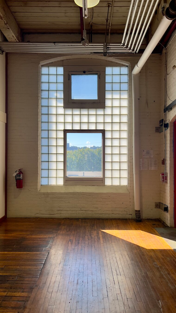 View toward a multi-paned window in an old industrial factory. Some of the panes are large, some are small, some transparent and some opaque. Pipes line the ceiling and there is a wooden floor. Beyond the main window you can see a landscape.