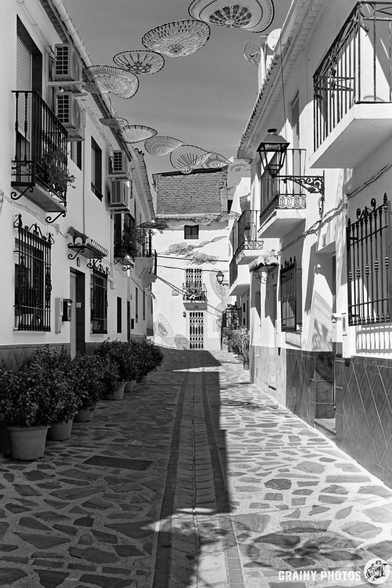 A narrow, cobbled street in Guájar-Fondón is lined with whitewashed buildings and balconies adorned with potted plants. Decorative parasols hang overhead, casting shadows across the black-and-white scene.