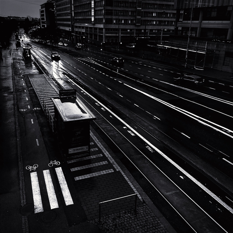 A dark, rainy street scene with road surface markings, a bus stop, and bike lanes, showcasing urban environment in monochrome.