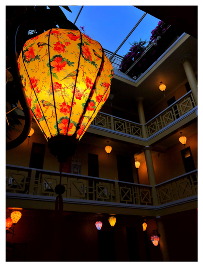 Photo of a hotel courtyard, taken at ground level and looking up past the railings of 2nd- and 3rd-floor balconies to a rectangle of clear evening sky. Suspended in the foreground at upper left is an illuminated Vietnamese lantern, with silk fabric in a design of colorful birds and orange flowers with green leaves against an orange background. Similar lanterns hang from the ceilings of the different levels of the hotel, glowing softly against the shadows. Past the uppermost level is a glimpse of shadowy plants with pink flowers on the rooftop.