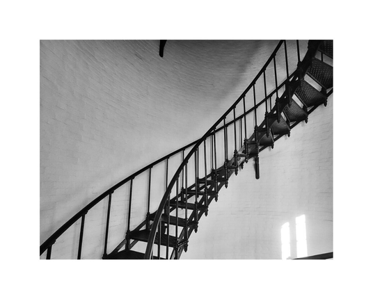 Curved metal lighthouse staircase spirals upward against a textured white brick wall in black and white, creating a dramatic, architectural composition. Light streams in.
