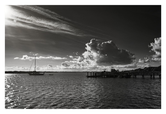 Black and white seascape with a sailboat on the left and a pier on the right. Large, dramatic clouds hover above, creating a tranquil, reflective mood.
