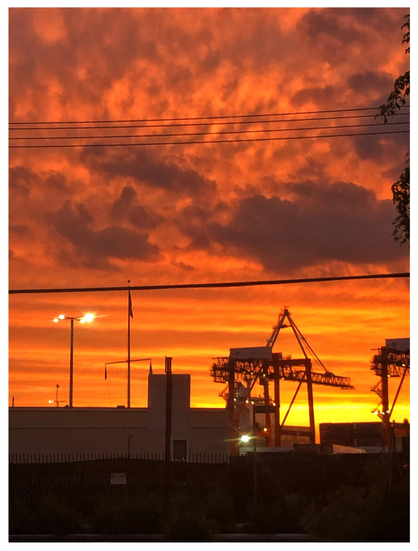 Photo of a dramatic orange sunset over the Brooklyn waterfront. In the foreground is a low building behind a fence, illuminated streetlights, some tree leaves at right frame, and a container crane with its boom lowered and extended, silhouetted against the sunset glow. Some power lines stretch across the sky from left to right frame. Dark clouds are backlit by orange light, which gradually turns yellow-gold at the horizon.