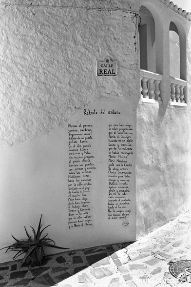 Black and white photo of a whitewashed building on Calle Real in Guájar-Fondón, with a poetry mural—handwritten in Spanish—on the wall and a potted plant at the corner of the cobbled street.
