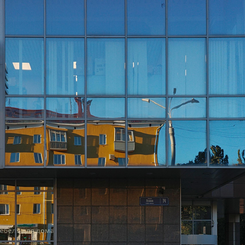The photo shows a building with large glass windows that reflect the sky and the surrounding environment. Other buildings can be seen in the reflection, including an orange building with balconies.