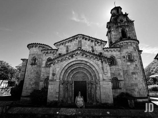 Fotografía en blanco y negro de una imponente iglesia de piedra de estilo románico. La fachada principal presenta un gran portal con múltiples arcos escalonados y profusas esculturas de figuras humanas y ornamentos tallados. A la derecha de la iglesia, un alto campanario de piedra se alza hacia el cielo, con el sol resplandeciendo justo detrás de su cima, creando un fuerte contraluz y un halo de luz. Frente a la entrada principal, una mujer joven, vestida con una camiseta clara y gafas de sol, posa de pie mirando hacia el frente. La iglesia tiene tejados a dos aguas de teja y varias secciones semicirculares. Alrededor se aprecian algunos árboles y vegetación, bajo un cielo parcialmente nublado.