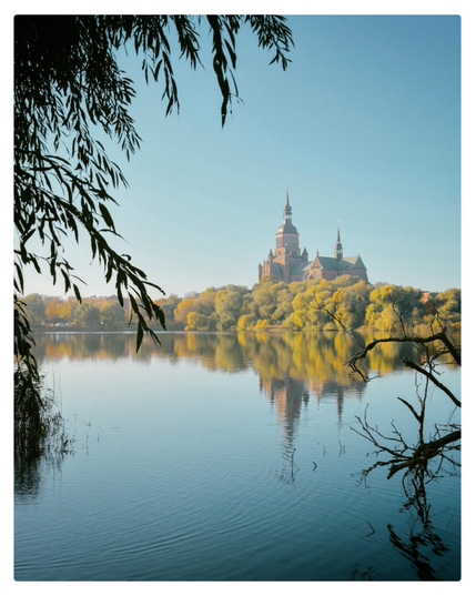 A tranquil lake scene with clear blue water reflecting a large historic brick church with tall spires in the distance. The shoreline is lined with autumn-colored trees in shades of yellow and green. Overhanging branches frame the image from the top and right, adding depth to the composition. The sky is clear and bright, enhancing the peaceful atmosphere.