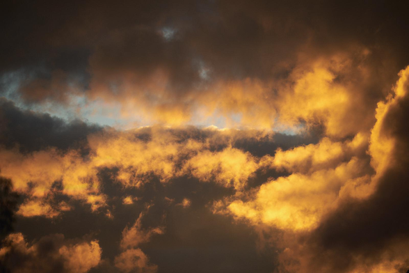 Golden, sunlit clouds billow across a dark, stormy sky, with small patches of blue peeking through the glowing gaps.