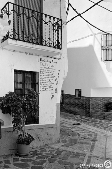 A narrow cobblestone street in Guájar-Fondón is lined with white buildings. One building features a small balcony, a potted plant, and a handwritten note beneath a poetry mural, with painted footprints leading towards a sunlit curved corner.
