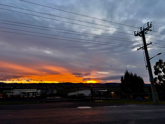 A blazing orange sunset at the horizon. Covered with grey clouds above. A telegraph pole, wires and industrial scene is dark. Almost in silhouette. 