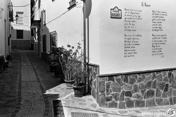 A narrow cobblestone alley in Guájar-Fondón, lined with potted plants. A tiled street sign reads Calle Rosario and a poetry mural titled El Rocío is written on a white wall. The scene is in black and white.