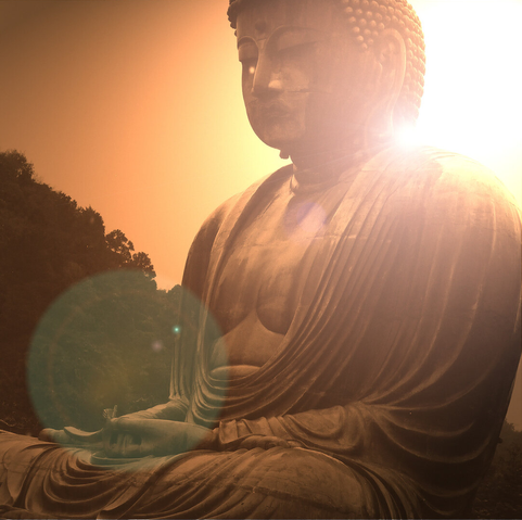 A color photo showing the Great Buddha statue at Kamakura, Japan. The figure is seated. Sunlight is peeking over the figure’s left shoulder, lending an orange hue to the scene.
