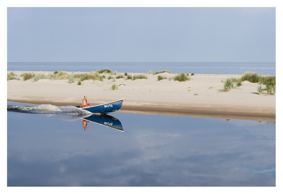 A person navigates a small motorboat across calm waters, creating gentle ripples that reflect the bright midday sky. The serene background features a sandy shoreline bordered by tall grasses, blending into a horizon of open water under soft clouds.