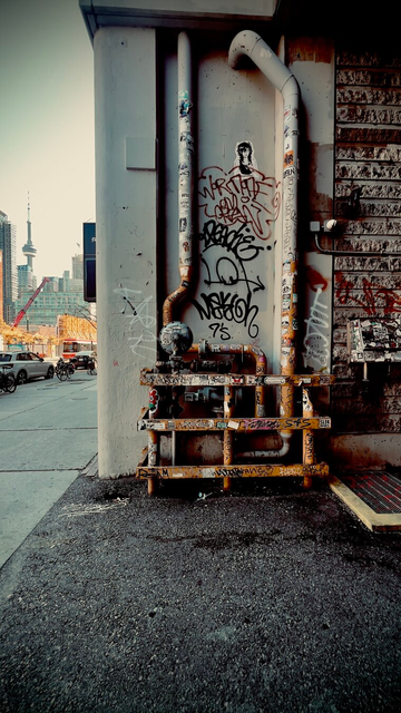 Rusted and graffitied pipes on the outside of a building. View shows a sliver of the street beyond that includes the CN Tower.