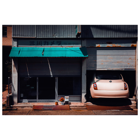 A color photo of an old storefront with a faded turquoise awning and partially closed metal shutters. The sign above reads “荒川カメラ” (Arakawa Camera). The left side of the shop is dark and empty, while on the right a pale pink car is parked halfway inside the garage. A small cactus and potted plant sit neatly on the ground out front, adding a touch of life to the otherwise still, sunlit scene.