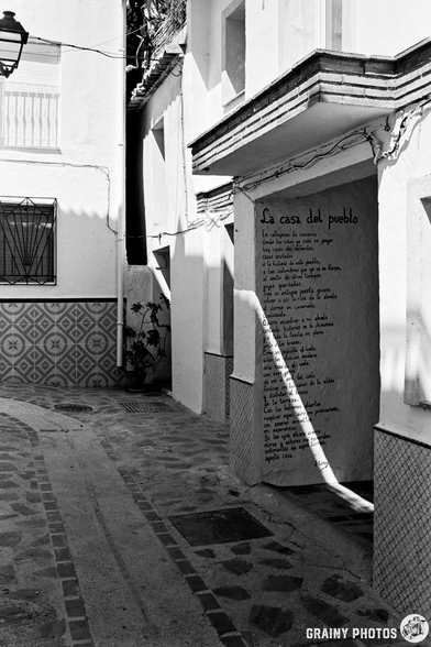 A narrow, sunlit alley with whitewashed walls in the Spanish village of Guájar-Fondón. One wall features a poetry mural titled La casa del pueblo. Decorative tiles and shadows add texture to the quiet, empty street.