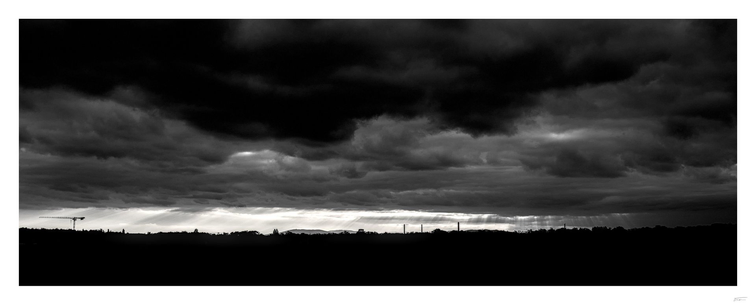 Dieses Foto zeigt eine dramatische Schwarz-Weiß-Aufnahme des Himmels über Wien. Der Himmel ist von dichten, bedrohlich wirkenden Wolkenformationen geprägt, die sich über die gesamte Bildbreite erstrecken. Am Horizont sind die Silhouetten von Bäumen, Gebäuden und einigen Schornsteinen zu erkennen. Durch die Wolkendecke brechen an einigen Stellen Lichtstrahlen, die dem Bild eine fast mystische Atmosphäre verleihen. Die Stimmung wirkt düster und eindrucksvoll.