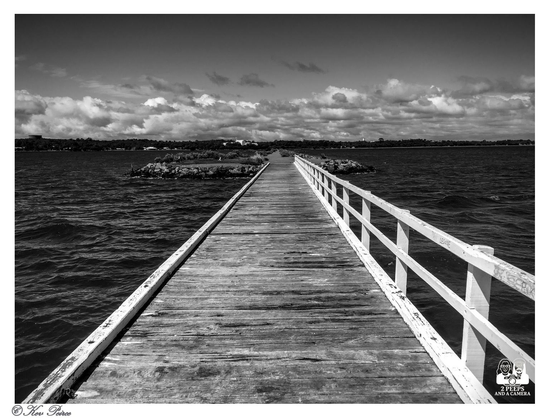 A striking black and white photograph of a wooden jetty extending straight out over a large body of water.

The weathered wooden planks of the jetty, framed by a white railing on the right side, create a strong leading line that draws the eye toward the distant shore and center.

The water is dark and choppy, and the sky is filled with dramatic, bright clouds that add texture and contrast to the scene.

The distant shoreline is dark, providing a strong horizontal line.

The bottom right corner is watermarked "© Kev Peirce."