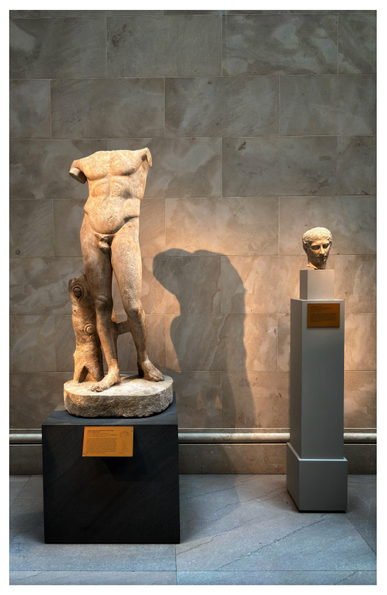 Photo of two marble Roman sculptures on pedestals at the Metropolitan Museum of Art in NYC. At left is an armless, headless statue of a male youth, with one leg braced against a sculpted tree stump. At right is the sculpted head of a man. The lighting throws the shadow of the headless statue onto the tiled marble wall between the two artifacts.

Info from the museum placards:

Marble statue of the Diadoumenos (youth tying a fillet around his head)
Roman, imperial period, 1st-2nd century A.D.
Copy of a Greek bronze statue of ca. 430 B.C. by Polykleitos

Marble head of a youth
Roman, probably Claudian period, ca. A.D. 41-54
Copy of a Greek bronze statue of ca. 450 B.C. attributed to Polykleitos 