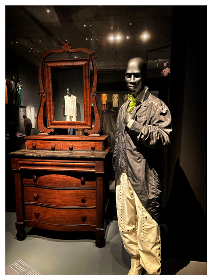 Photo of a bald, dark-toned, male mannequin wearing a long gray bomber jacket over a striped collared shirt, green necktie, and light-colored pants studded with rivets. Beside it is an antique bureau with brown wooden drawers topped by a framed rectangular mirror, which reflects the image of a similar mannequin in a sleeveless white top. In the background are scattered displays of more mannequins in various outfits, under a dark ceiling dotted with museum lights.

Museum placard info for the main mannequin:

denzilpatrick (British, founded 2021)
Daniel Gayle (British, b. 1983)
James Bosley (British, b. 1983)
Ensemble, autumn/winter 2024-25
Tailed bomber jacket of gray synthetic twill; trousers of ivory cotton tumbled velvet studded with rivets of bronze metal; shirt of gold-and-ivory striped cotton plain weave; tie of green nylon plain weave; spats of ivory cotton tumbled velvet and bronze metal
