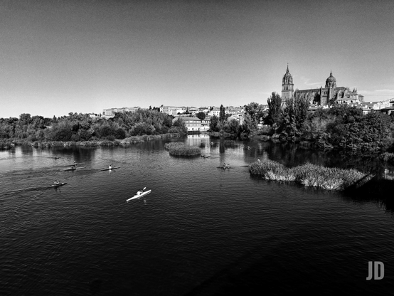 Una fotografía en blanco y negro muestra una vista panorámica de un río ancho y tranquilo. En primer plano y en el centro, varias personas reman en kayaks o canoas; un kayak blanco se destaca en la parte inferior derecha, mientras otros se ven más lejos a la izquierda. El río está salpicado de islas pequeñas con vegetación densa, como juncos. Las orillas están bordeadas por una exuberante vegetación de árboles. Al fondo, sobre una colina o elevación, se extiende una ciudad, dominada por una majestuosa catedral con grandes torres y cúpulas. El cielo es claro y luminoso, sugiriendo un día soleado.