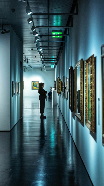 Das Bild zeigt eine Person, die in einem langen, hell beleuchteten Flur einer Kunstgalerie oder eines Museums steht. Die Person blickt auf die Gemälde, die in goldenen Rahmen an der rechten Wand hängen. Der Flur hat einen glänzenden Boden, der die Lichter und die Umgebung reflektiert. Am Ende des Flurs sitzt ein Museumswächter auf einem Stuhl.