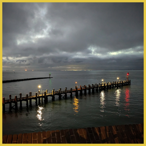 A broad river in first morninglight, heavy clouds in the sky. 2 piers in front, far away the other bank. Some lights brightens the riverwater