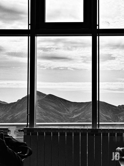 Una fotografía en blanco y negro muestra una vista panorámica de montañas a través de una ventana. El marco oscuro de la ventana divide la imagen en varias secciones, con una cruz prominente en el centro superior. Más allá del cristal, se aprecian grandes montañas con picos y laderas detalladas, extendiéndose hasta el horizonte. El cielo está cubierto de nubes dispersas, creando un contraste marcado con las siluetas montañosas. En la parte inferior de la imagen, se ve un radiador de calefacción de color oscuro que se extiende horizontalmente bajo la ventana, y a la izquierda, una forma oscura y borrosa sugiere la presencia de una persona o un objeto en primer plano.