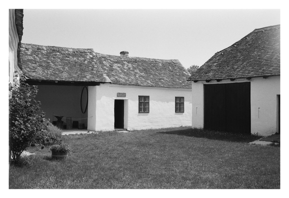 Blick in den Innenhof des Ausstellungsbauernhofes im Museumsdorf Niedersulz. Ein organisch geschwungenes Schindeldach ruht auf kalkweißen Wänden des Mauerwerks.