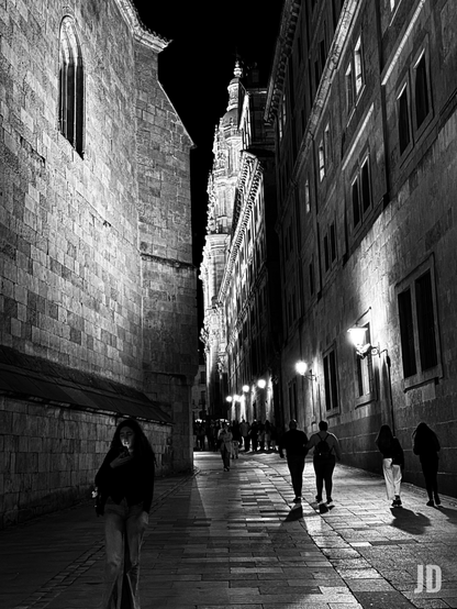 Fotografía en blanco y negro de una calle estrecha y antigua por la noche, flanqueada por edificios históricos. A la izquierda, se alza una imponente pared de piedra de un edificio antiguo, posiblemente una catedral o iglesia, con una ventana arqueada y alta visible en la parte superior. A la derecha, otro edificio de piedra, también alto, con múltiples ventanas oscuras y farolas ornamentadas que proyectan círculos de luz sobre el suelo. Al fondo, entre ambos edificios, se vislumbra la silueta iluminada de una torre o campanario con detalles arquitectónicos intrincados. En primer plano y ligeramente a la izquierda, una mujer joven con cabello largo y oscuro camina hacia adelante, mirando la pantalla iluminada de su teléfono móvil, que ilumina su rostro. Varias personas más se ven caminando por la calle adoquinada, algunas en parejas o pequeños grupos, creando sombras alargadas bajo la luz de las farolas. La iluminación artificial crea fuertes contrastes entre la luz y la oscuridad, acentuando la textura de la piedra y dando una atmósfera dramática y misteriosa a la escena nocturna."