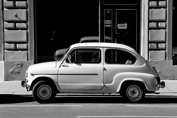 The black and white image shows a small vintage car parked neatly along a city street in front of a building with textured walls and recessed panels. The car’s smooth, rounded shape and simple details evoke a classic mid-20th-century design, and sunlight casts sharp shadows that emphasize its contours.