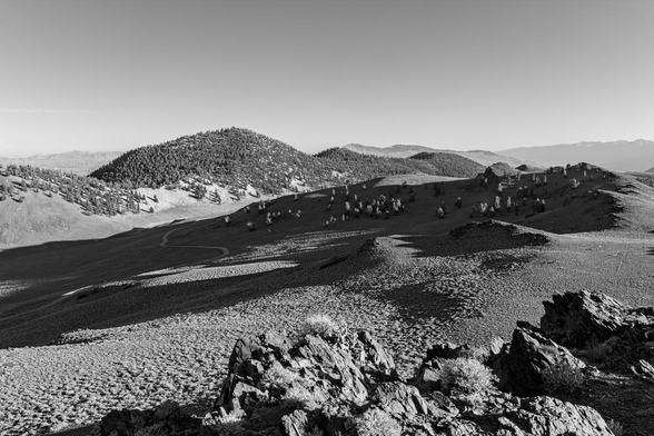 A black and white landscape photo from a mountain summit looking out over a sagebrush covered landscape with a  few trees dotted on the slope of a hill from the center to the right and a further distant series of three hills thickly covered in trees. There are some very jagged rocks in the lower right foreground. A heavy shadow cuts across from lower left to center-right.