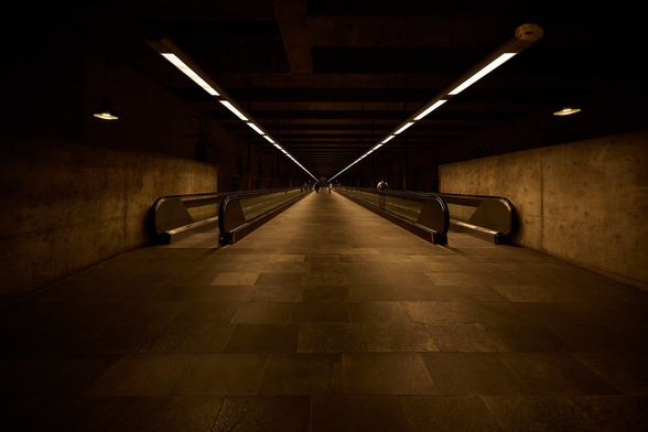 The photo shows a long pedestrian tunnel of a metro station. The photo is very symmetrical, the lights on the ceiling and the straight escalators left and right are all running towards a vanishing point in the center.