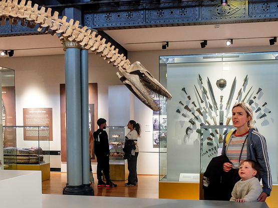 A young woman and a child are looking at a dinosaur skeleton that seems to be looking back at them. At the back of the room, a man and a woman are also talking face to face.