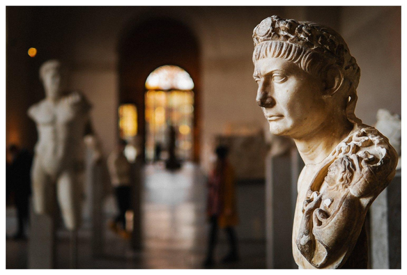Blick in ein Museum. Links die Büste eines Romers, im Hintergrund weitere Statuen, Menschen, ein lichtdurchfluteter Durchgang.
