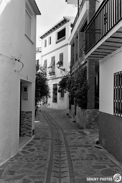 Narrow cobblestone street in Guájar-Fondón, lined with white buildings, balconies, and shuttered windows; plants and lanterns add charm to the quiet, sunlit scene.