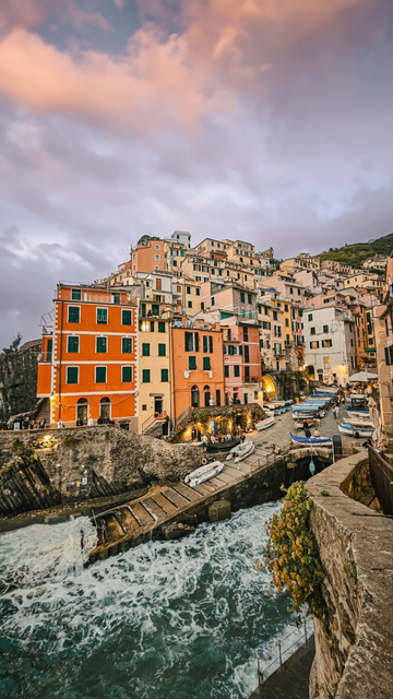 Colorful cliffside buildings of Riomaggiore, one of Italy’s Cinque Terre villages, rise above a narrow harbor filled with small boats and crashing turquoise waves. The sunset sky glows in soft pink and purple tones, casting warm light over the pastel facades and stone walkway leading to the sea.